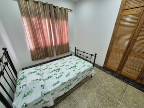 A single bed with a metal frame, a white bedspread with green leaf patterns, a wooden door, and a window with brown curtains.