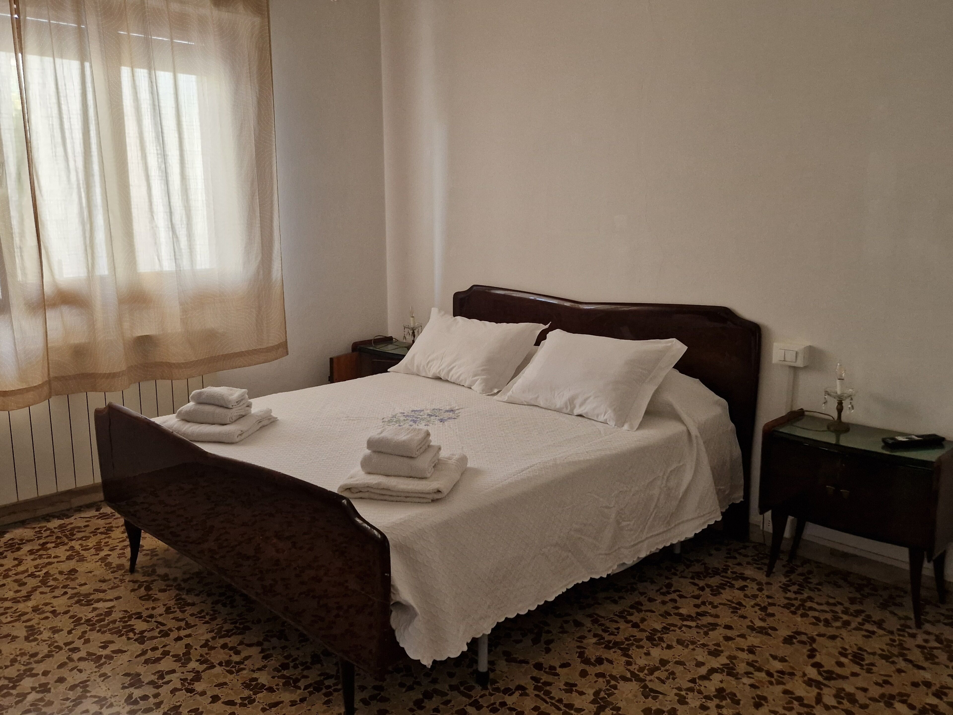 Un lit bien fait, avec des oreillers et des serviettes pliées, une table de chevet avec une lampe et une chaise dans une pièce dont le sol est recouvert d’un motif décoratif.