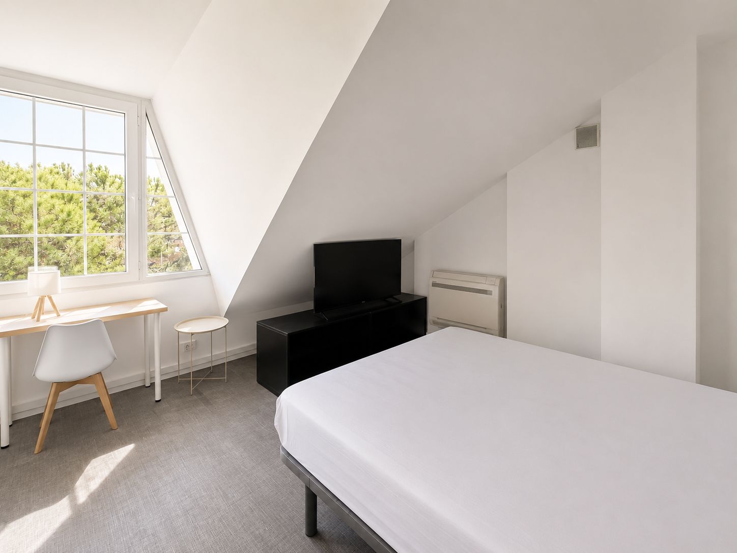 A modern bedroom with a sloped ceiling, a bed, a desk with a chair, a television, and a window with a view of trees.