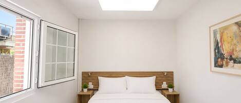 A bedroom with a bed, two bedside tables, a window, a ceiling light, and a painting on the wall.