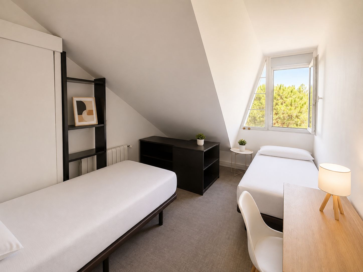A small, modern bedroom with a sloped ceiling, two single beds, a desk, a chair, a small table with a lamp, and a window with a view of trees.