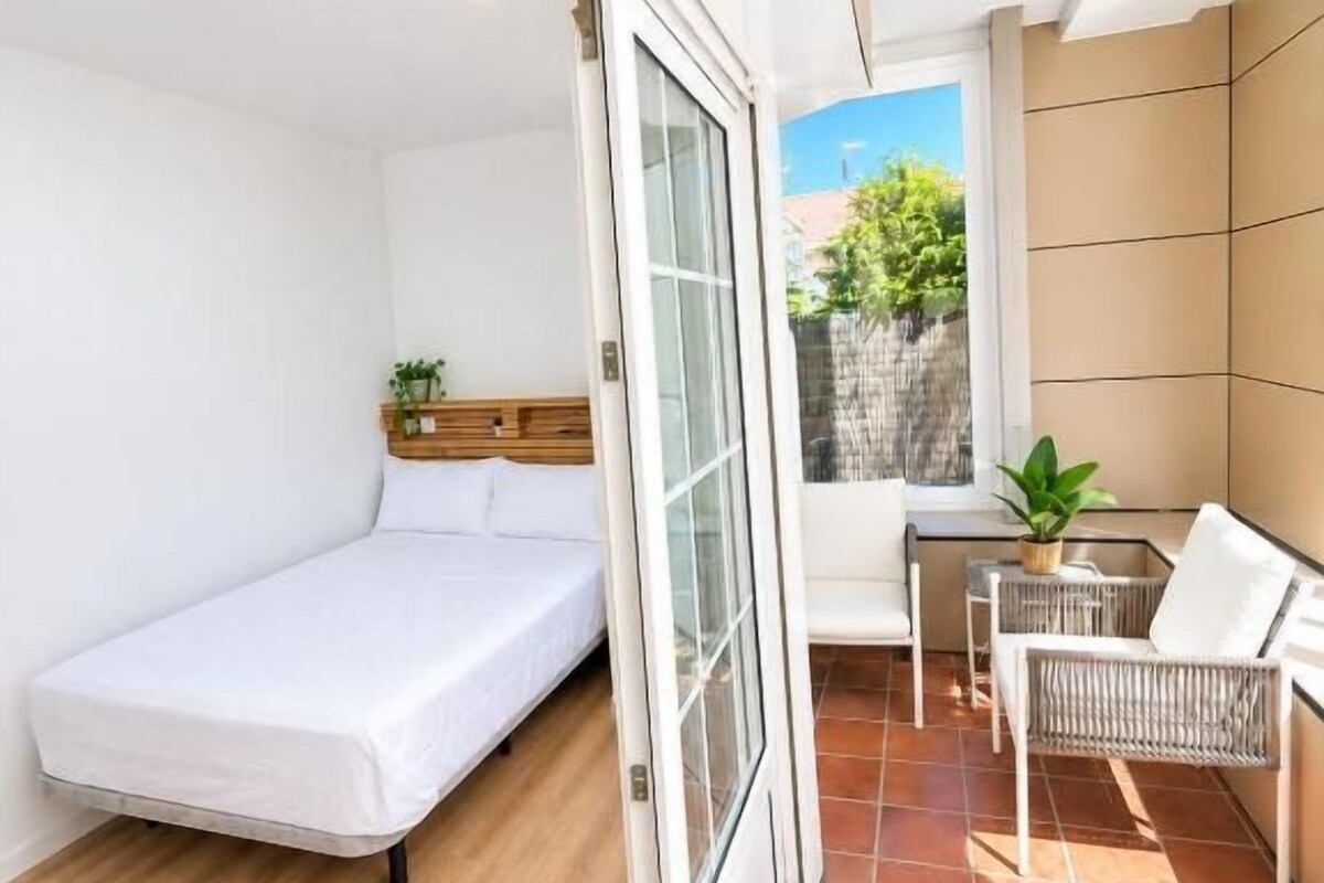 Habitación pequeña con cama, puerta corredera de vidrio a un área exterior, una mesita con una planta y una silla.