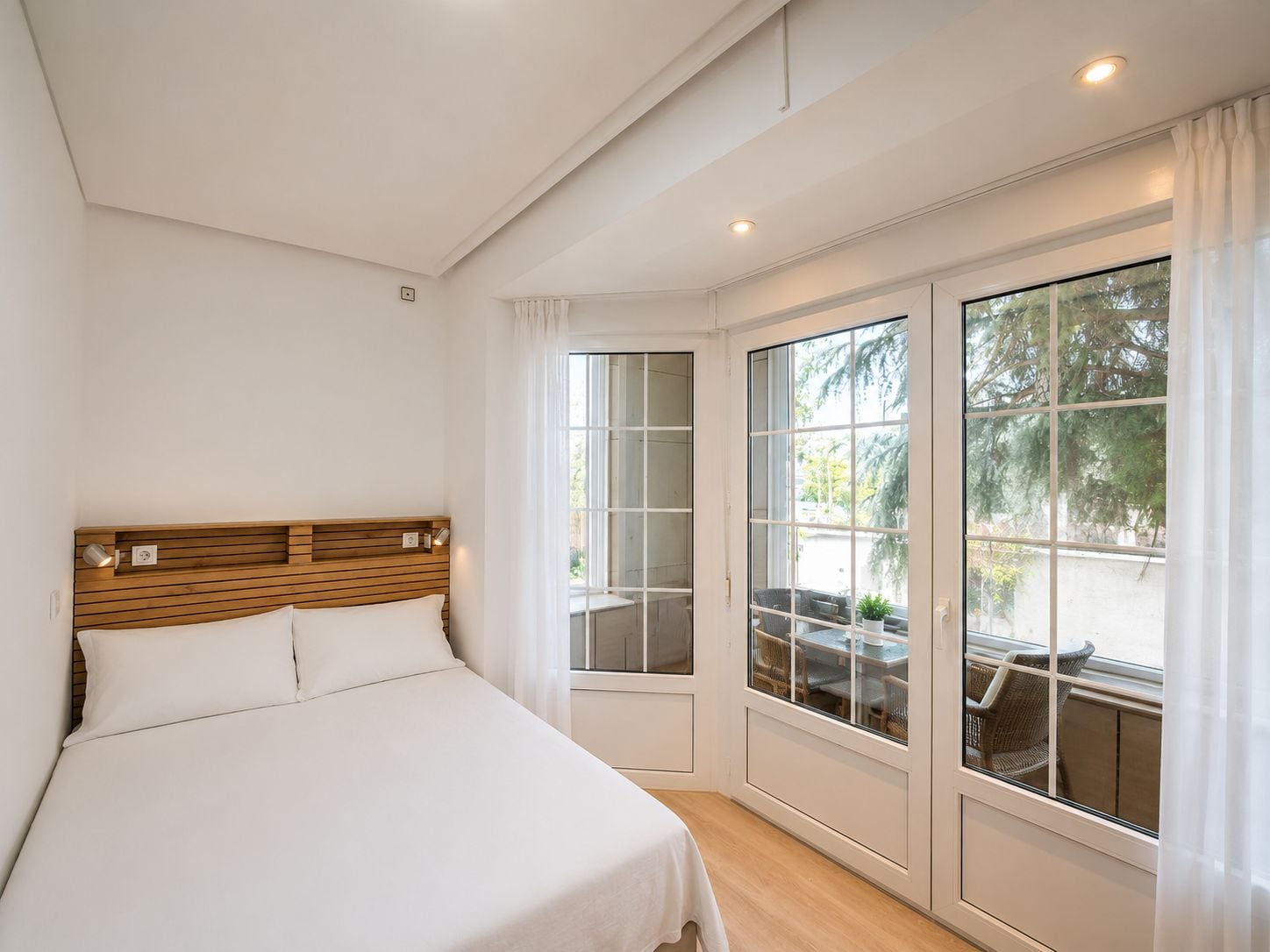 Een slaapkamer met een groot bed, een houten hoofdbord en uitzicht op tuinmeubels door een raam.