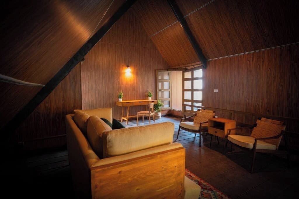 Quarto aconchegante com teto inclinado, paredes de madeira, um sofá, cadeiras, uma mesinha e uma janela com vista.