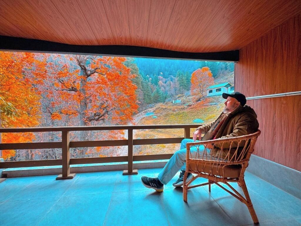 A person seated on a wicker chair in a room with a view of autumn trees through a large window.