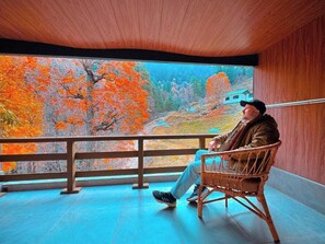 Une personne est assise sur une chaise en osier, dans une pièce d’où l’on peut admirer, à travers une grande fenêtre, les arbres aux couleurs automnales.