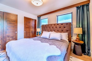 A large bed with a tufted headboard, two bedside lamps, and a view of the sky through windows. - Upstay Lake Dillon Basecamp Mountain Trails (Silverthorne)