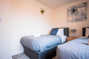 Une chambre d’hôtel avec deux lits, une table de chevet avec une lampe, une plante fixée au mur et un grand tableau abstrait.