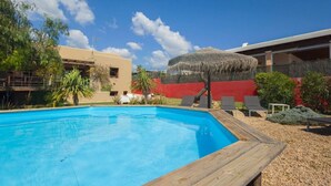 A swimming pool with a wooden deck, surrounded by a red wall and a building with a thatched roof. - Classic Ibiza Villa · Pool + Garden + 3 Bedrooms (Balearic Islands)