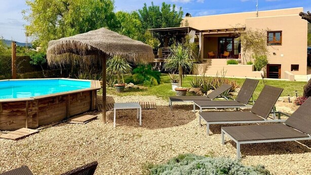 A pool area with lounge chairs, a thatched umbrella, and a building with a modern design. - Classic Ibiza Villa · Pool + Garden + 3 Bedrooms (Balearic Islands)