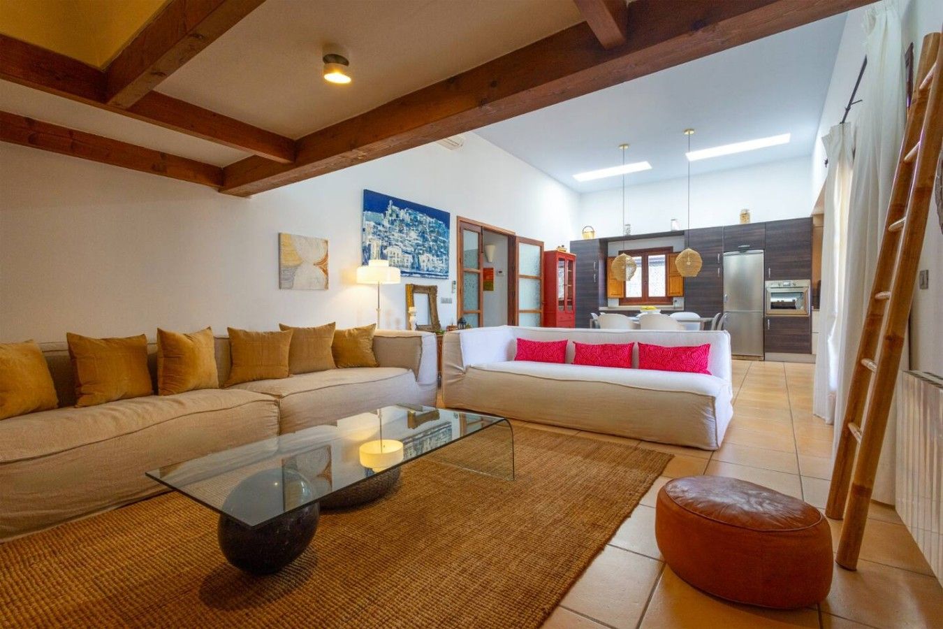 A modern living room with a glass coffee table, sofas with cushions, a kitchen area with cabinets and appliances, and a wooden ceiling structure.