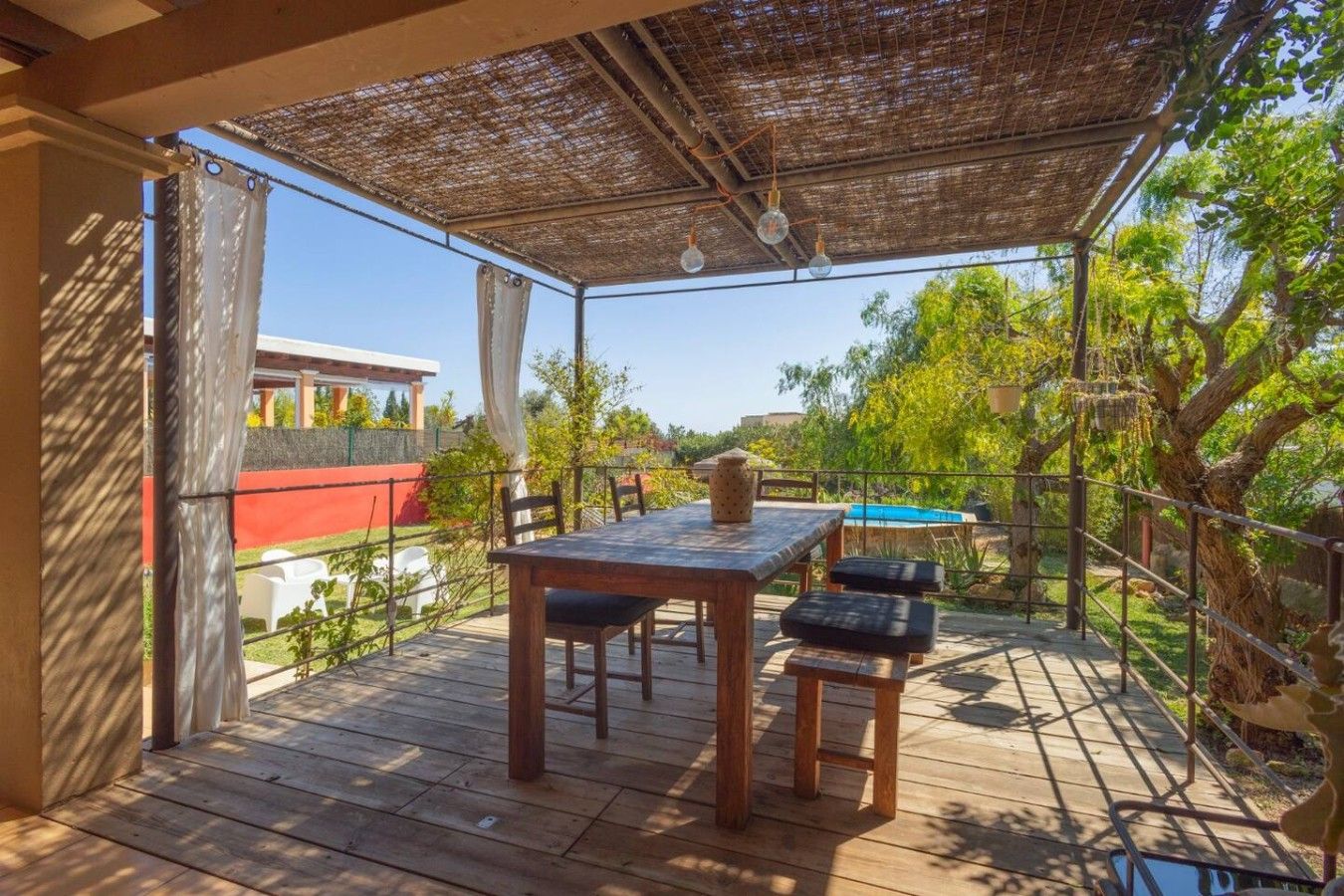 Eine Terrassenanlage aus Holz mit Tisch und Stühlen, ein überdachter Bereich mit hängenden Lichtern und Blick auf einen Swimmingpool und Bäume.