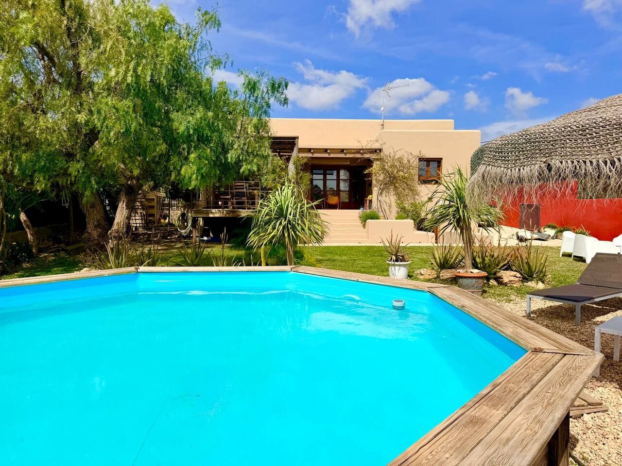 Une piscine dotée d’une terrasse en bois, entourée de verdure et bordée d’un bâtiment au toit de chaume.