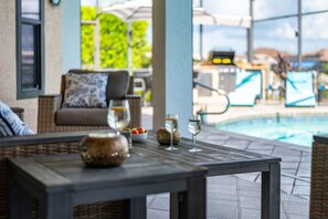A poolside area with a table, two glasses of water, a bowl of fruit, and a wicker chair with a cushion. - Villa Glasswater - Lake View, Kajaks & Dock (Cape Coral)