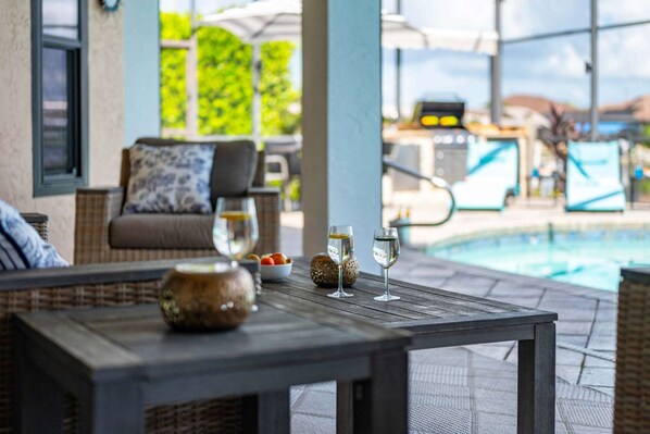 A poolside area with a table, two glasses of water, a bowl of fruit, and a wicker chair with a cushion. - Villa Glasswater - Lake View, Kajaks & Dock (Cape Coral)