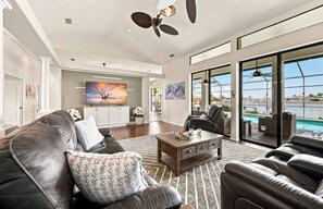 A spacious living room with a large window, a ceiling fan, and a view of a swimming pool. - Villa Glasswater - Lake View, Kajaks & Dock (Cape Coral)