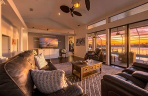 A spacious living room with a large sofa, a coffee table, and a view of a sunset through large windows. - Villa Glasswater - Lake View, Kajaks & Dock (Cape Coral)
