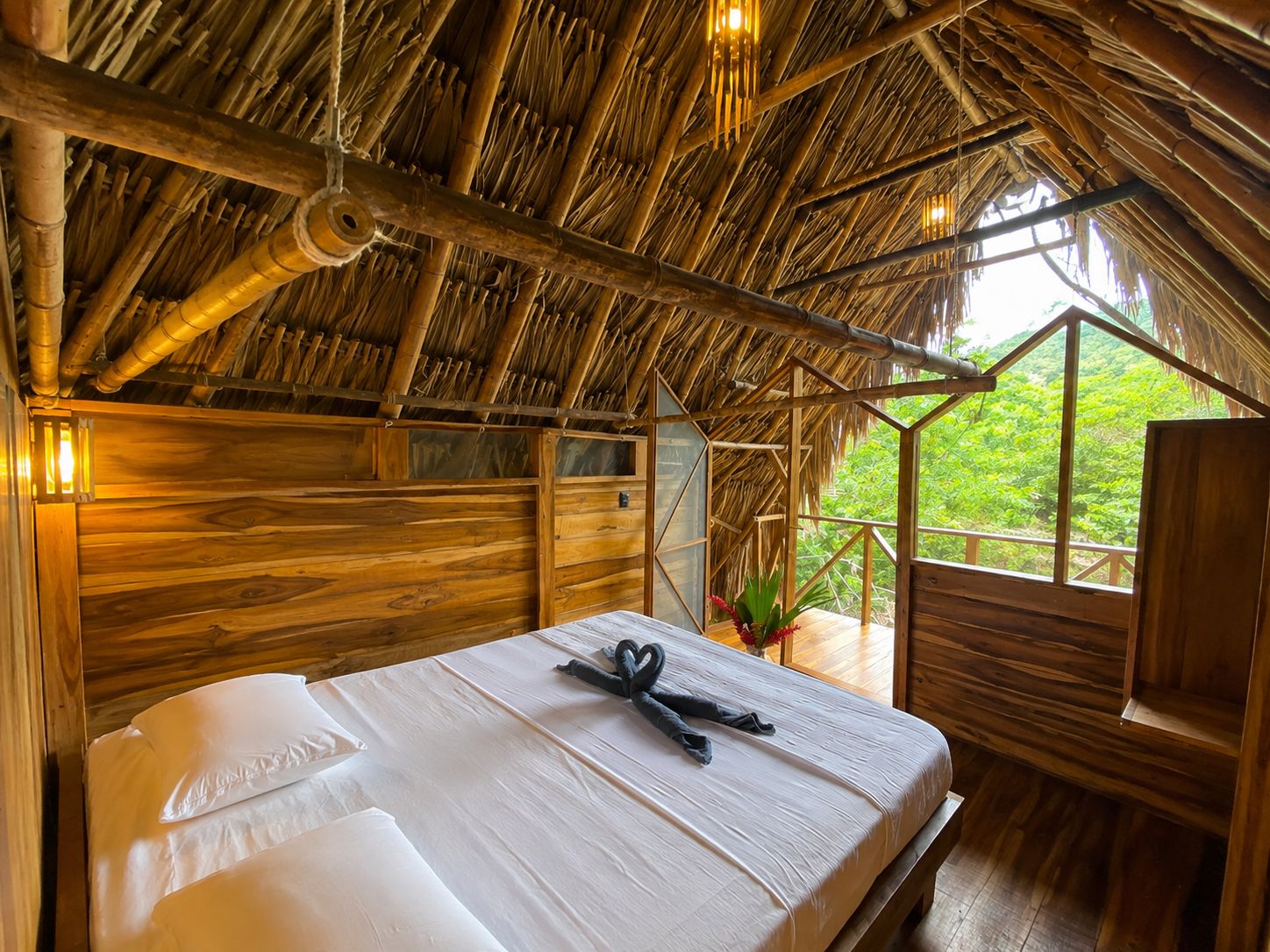 Ein Zimmer mit Strohdach, Holzwänden, einem Bett mit weißer Bettwäsche und einem Fenster mit Blick auf Grün.