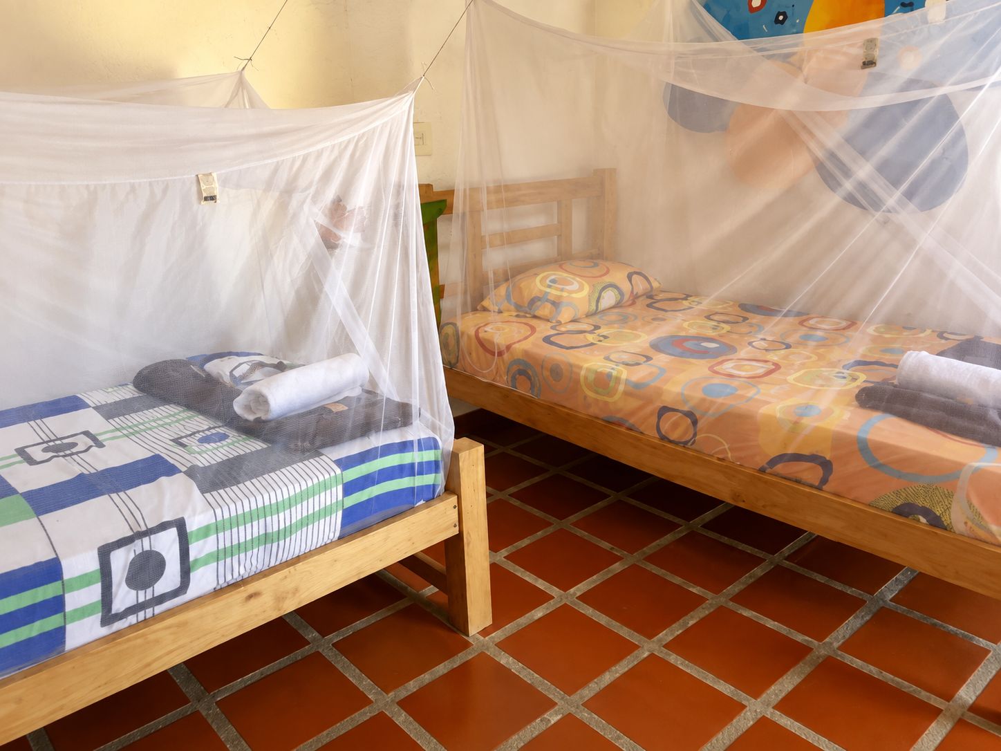 Dos camas de madera con mosquiteros, una con almohada a rayas y otra con almohada estampada, sobre un piso de baldosas.