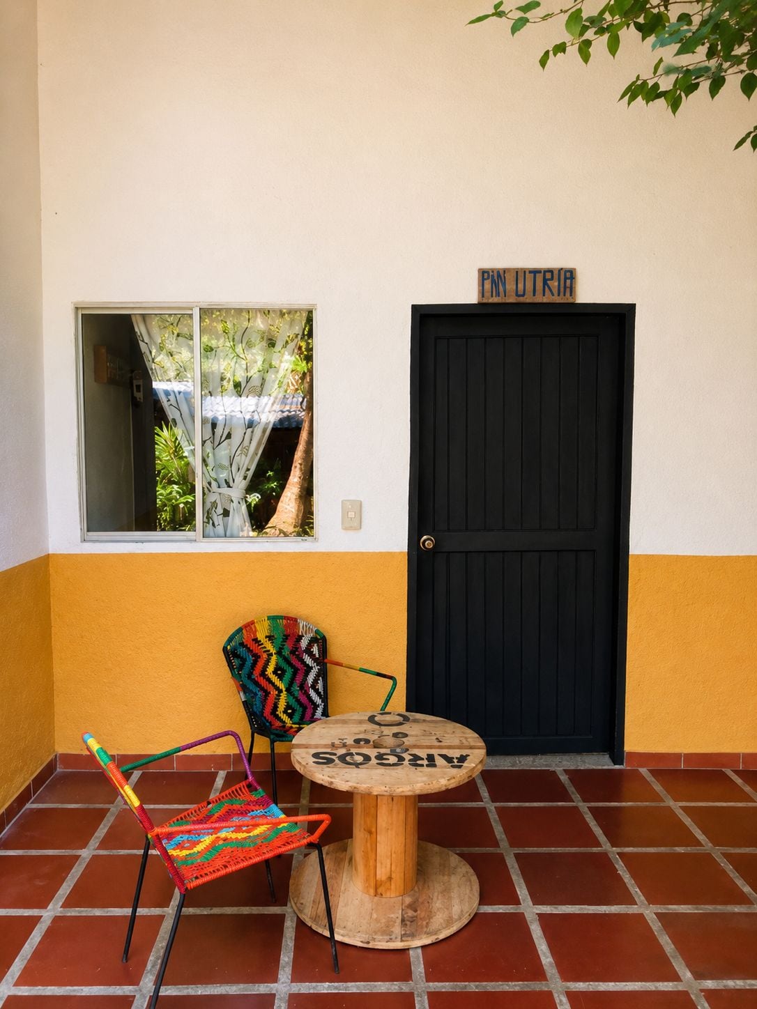 Una habitación con una puerta negra, una ventana que da a zonas verdes, un sillón de mimbre colorido y una pequeña mesa de madera.