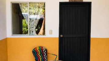 Una habitación con una puerta negra, una ventana que da a zonas verdes, un sillón de mimbre colorido y una pequeña mesa de madera.