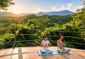 Dos personas practicando yoga en colchonetas al aire libre, con una vista panorámica de un bosque y montañas al fondo.