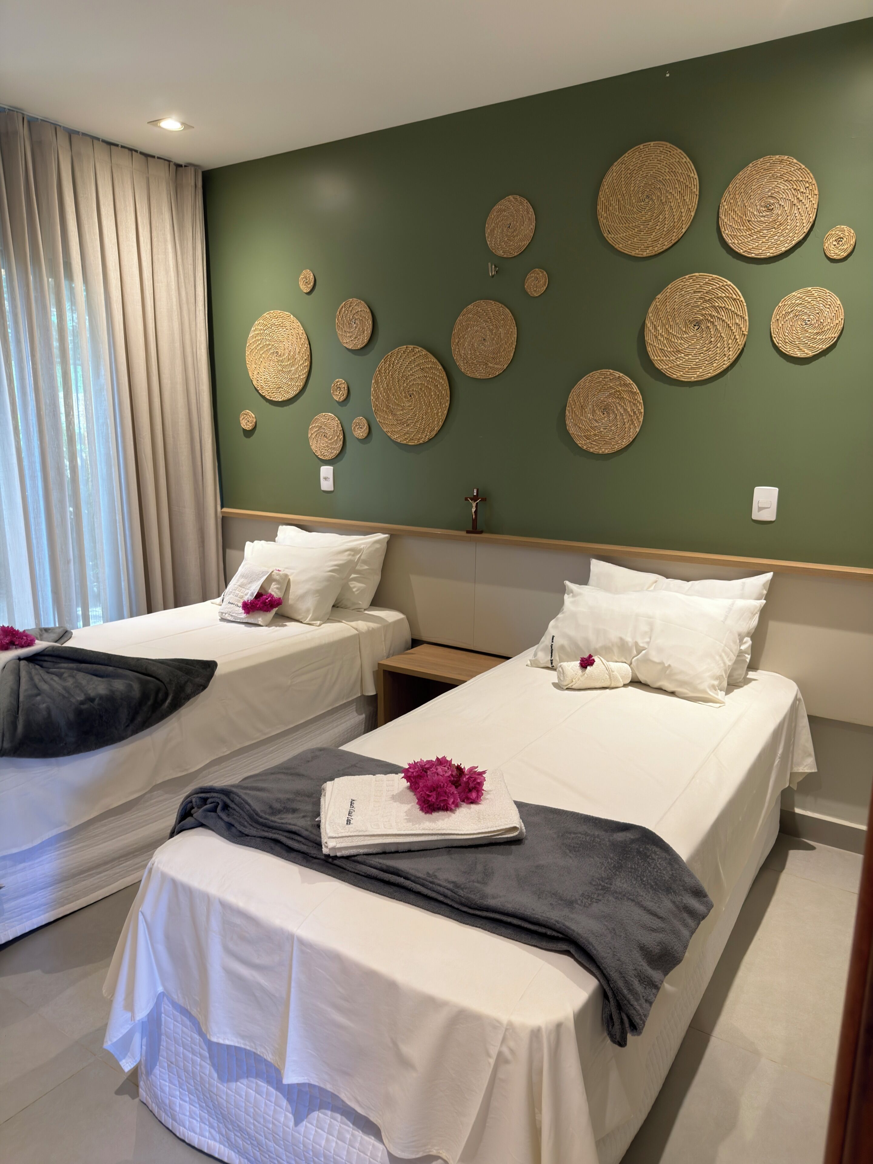 Quarto de hotel com duas camas, parede verde com arte em tecido, janela com cortinas e mesa de cabeceira com decoração em cruz.