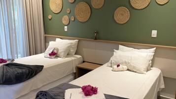 Quarto de hotel com duas camas, parede verde com arte em tecido, janela com cortinas e mesa de cabeceira com decoração em cruz.