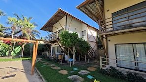 A two-story building with a wooden staircase and balcony, surrounded by greenery and palm trees. - Condomínio Amaré (Cajueiro da Praia)