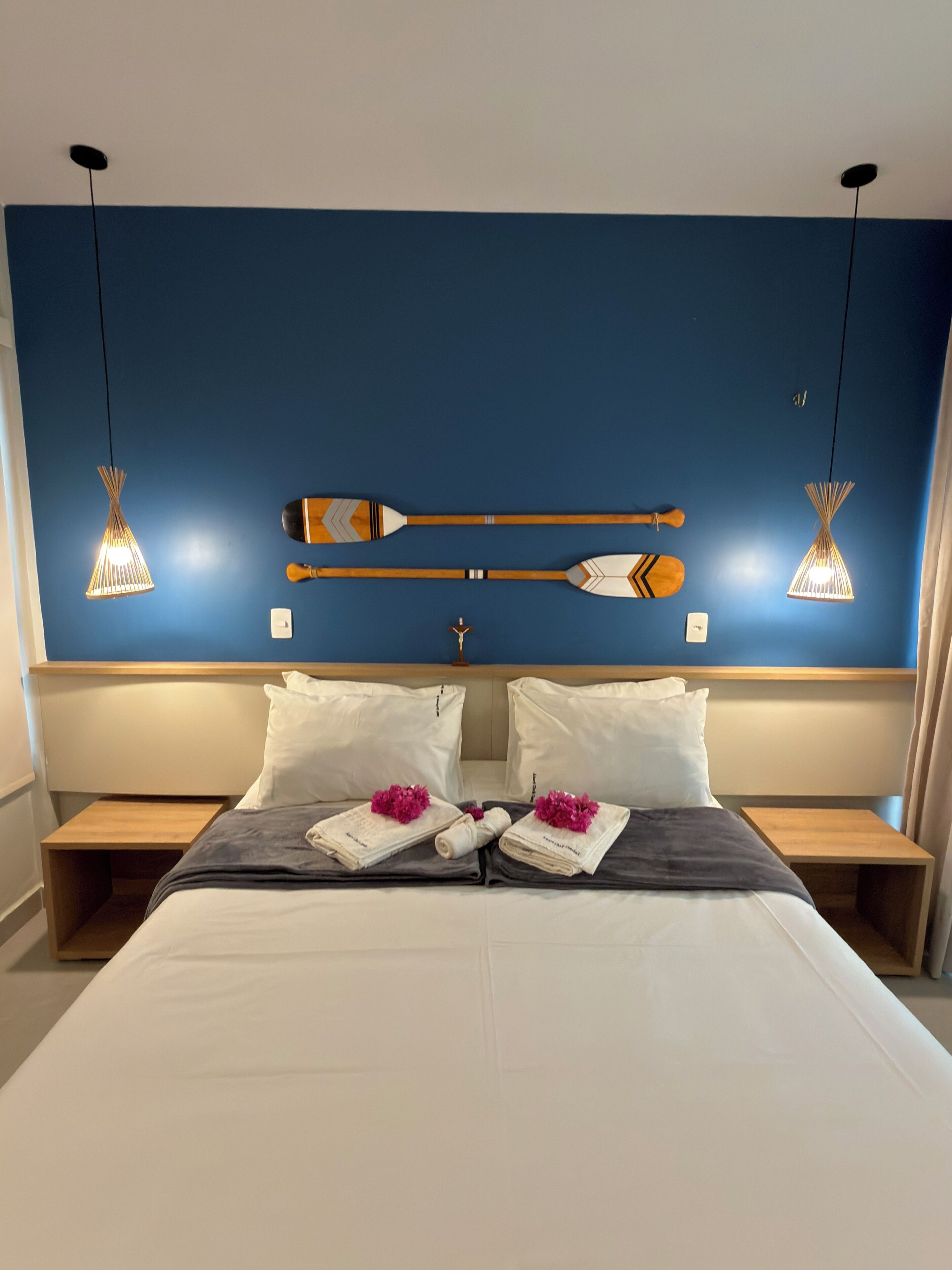 Una habitación de hotel con una cama grande, dos mesitas de noche, un cabecero con detalles decorativos e iluminación colgante.