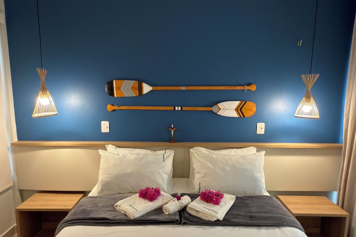 Una habitación de hotel con una cama grande, dos mesitas de noche, un cabecero con detalles decorativos e iluminación colgante.
