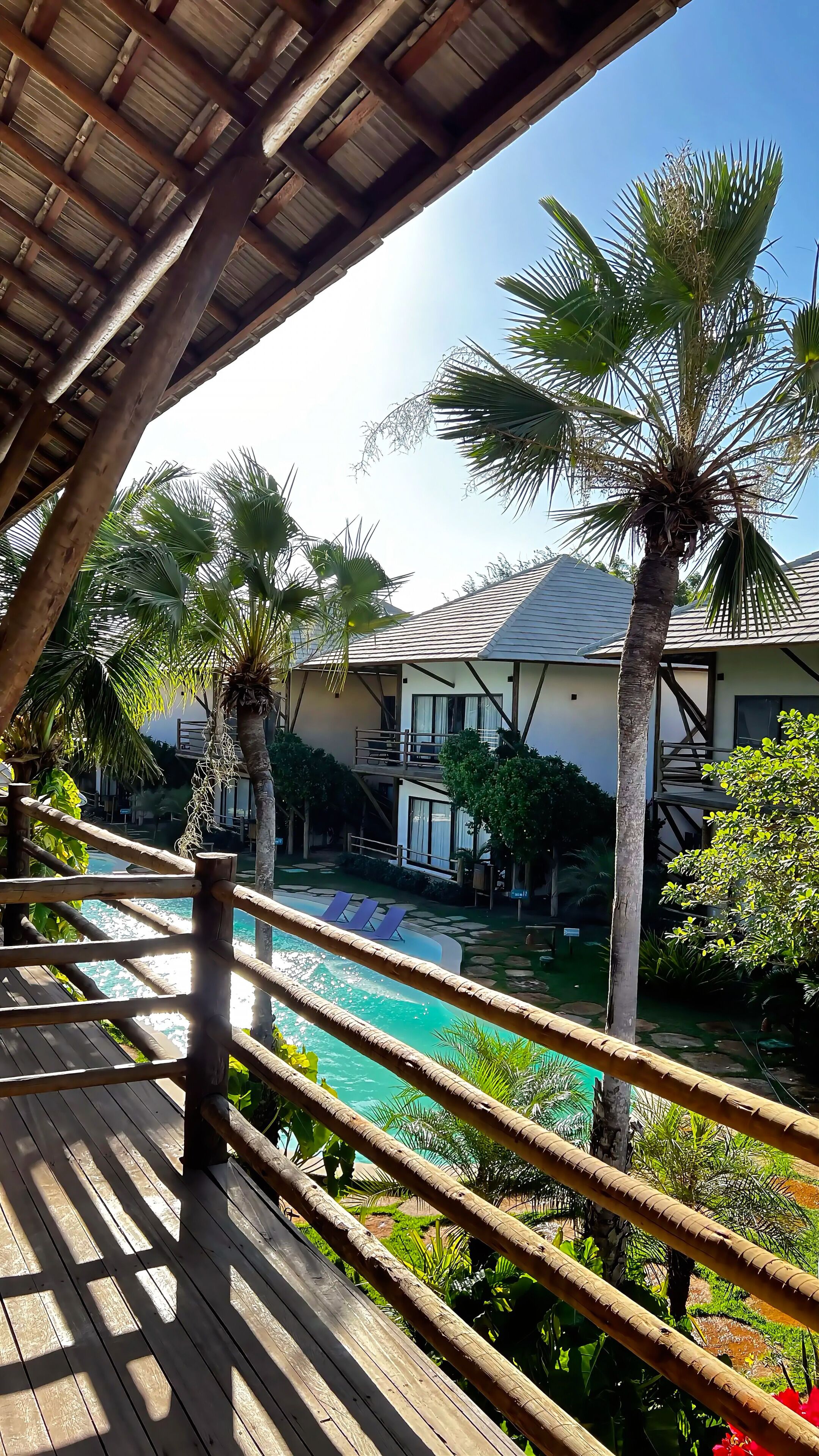 A wooden deck overlooking a swimming pool with palm trees and a building with a sloped roof.
