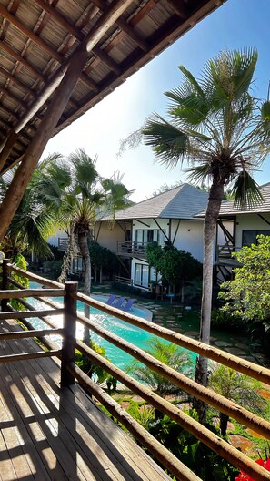 A wooden deck overlooking a swimming pool with palm trees and a building with a sloped roof. - Condomínio Amaré (Cajueiro da Praia)