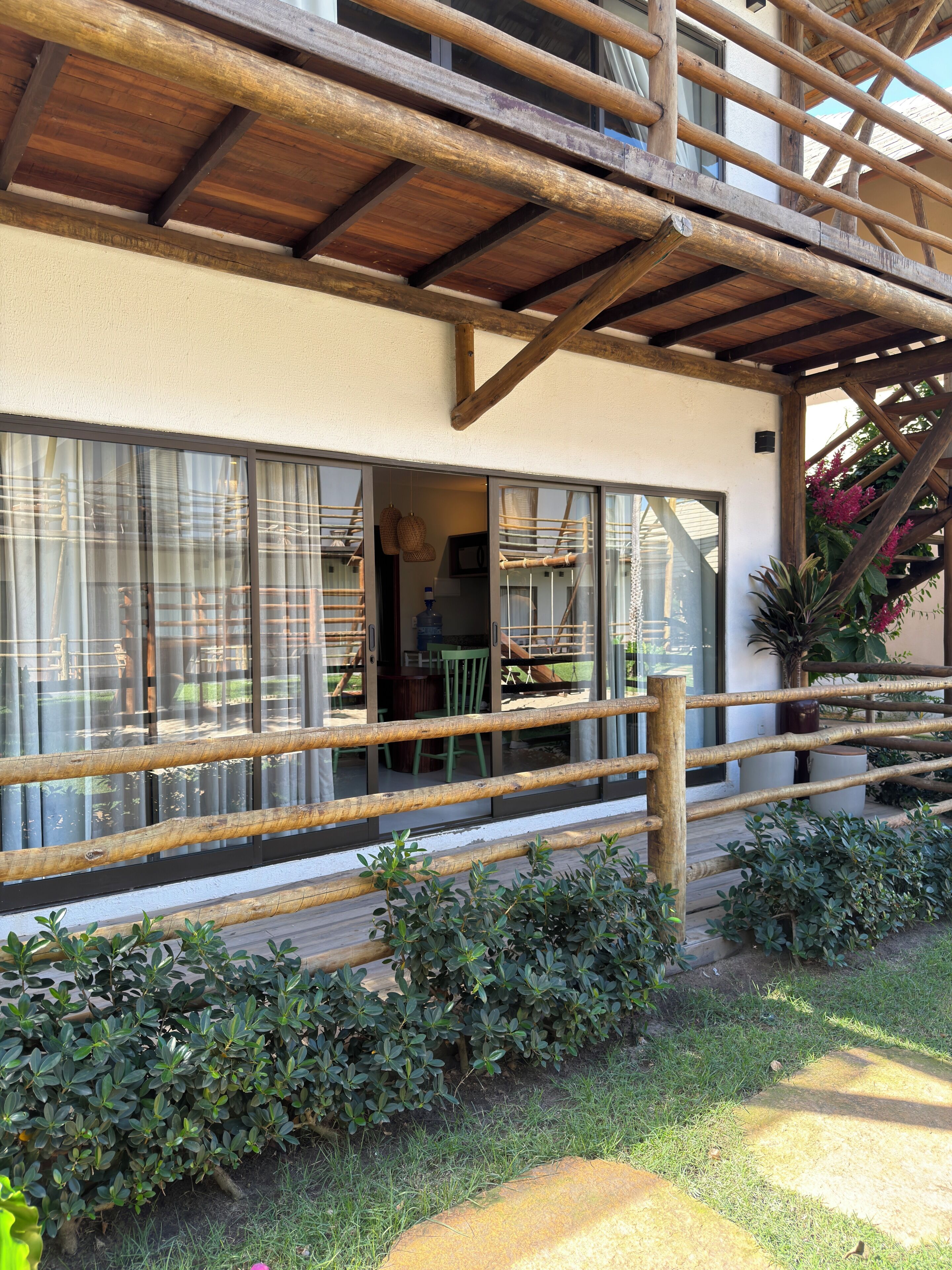 A wooden deck with a railing, a covered patio area, and a view of the interior through large windows.