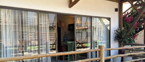 Um deck de madeira com guarda-corpo, uma área de varanda coberta e vista do interior através de grandes janelas.
