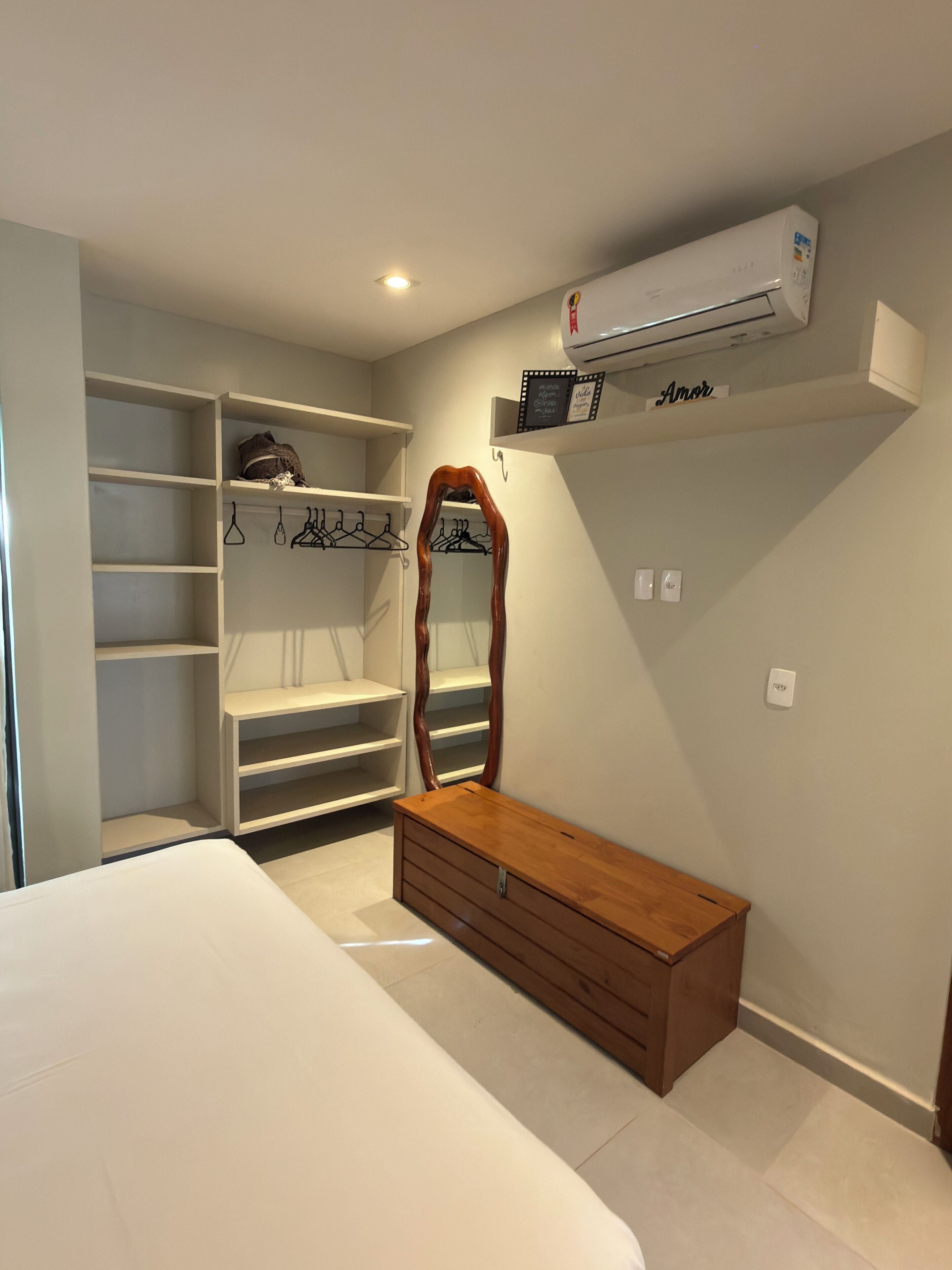 Ein Schlafzimmer mit einem Bett, einer Holzkommode, einem Schrank mit Regalen, einem Spiegel und einer Klimaanlage.