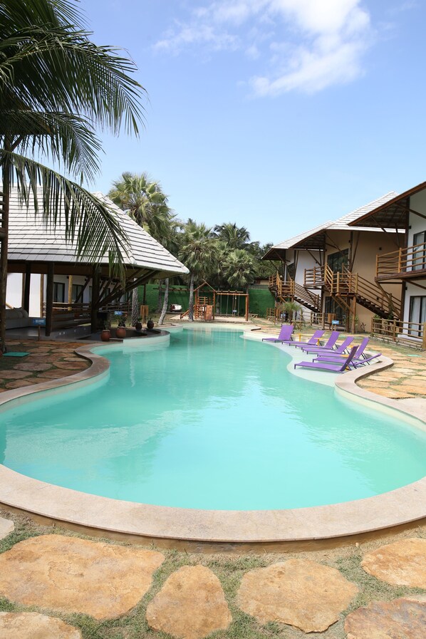 A swimming pool with a curved edge, surrounded by stone tiles and grass. There are purple lounge chairs and a wooden staircase leading to a building.