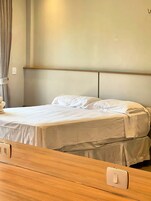 Cama com cabeceira de madeira, colcha branca e mesa de cabeceira com interruptor de luz.