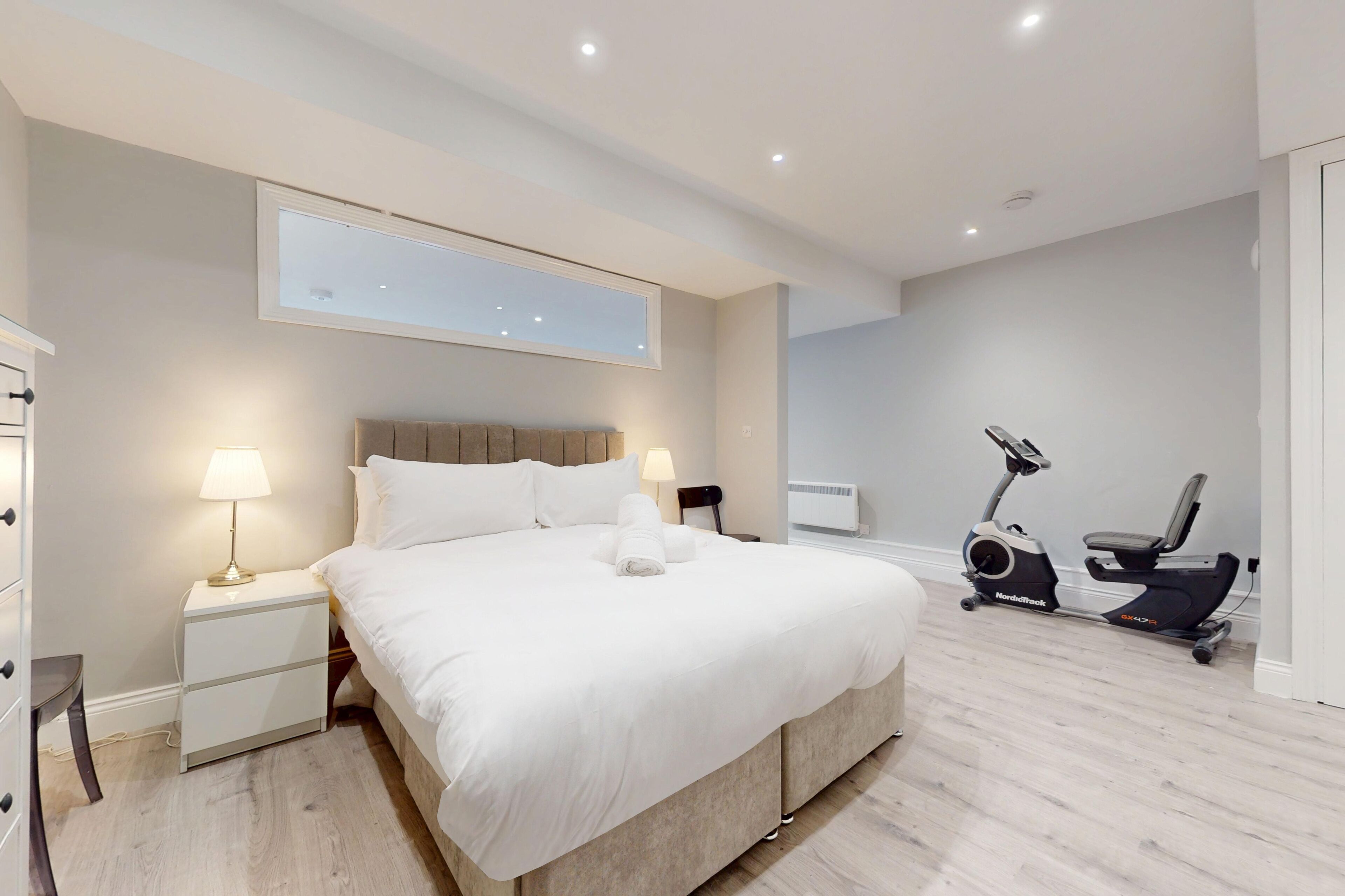A modern hotel room with a large bed, bedside lamps, a desk, a chair, and a stationary bike.