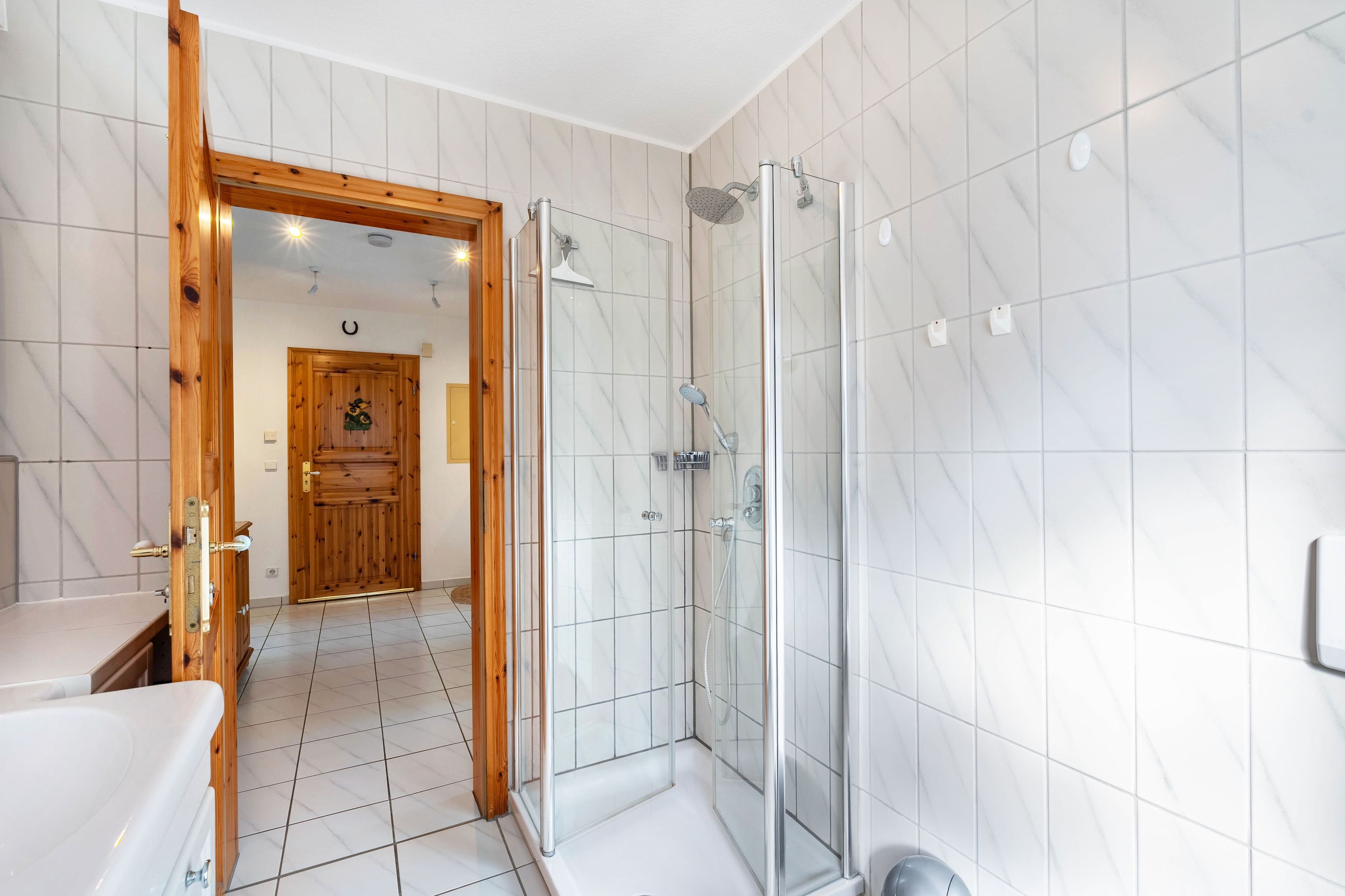 Un baño moderno con cabina de ducha de vidrio, puerta de madera y suelo de baldosas blancas.