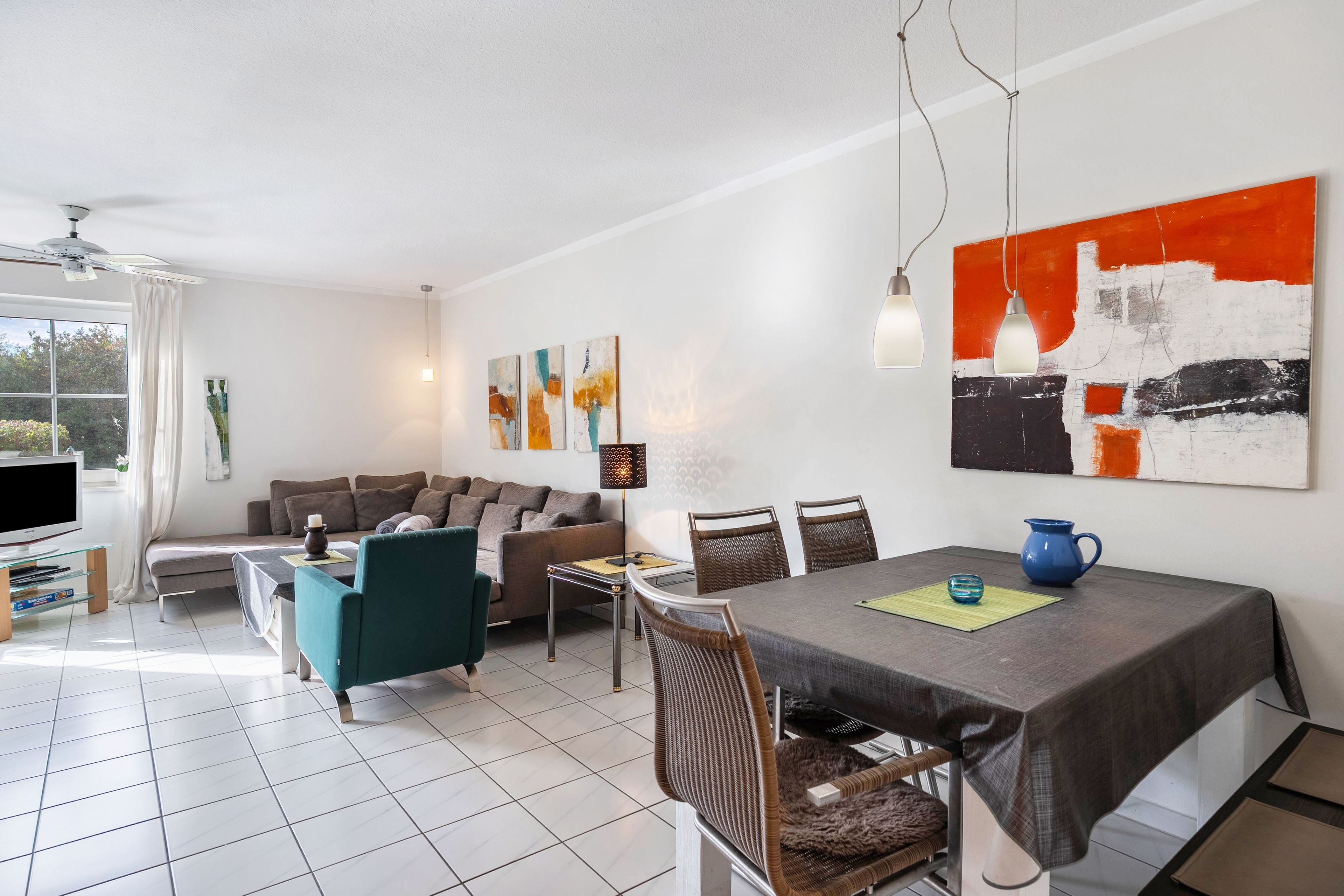 Ein modernes Wohnzimmer mit einer Couch, einem Esstisch und Stühlen. Es gibt einen Fernseher, einen Deckenventilator und Kunstwerke an den Wänden.