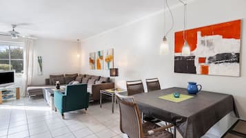 Ein modernes Wohnzimmer mit einer Couch, einem Esstisch und Stühlen. Es gibt einen Fernseher, einen Deckenventilator und Kunstwerke an den Wänden.