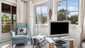 A living room with a blue armchair, a flat-screen TV on a glass table, and a view of greenery outside.