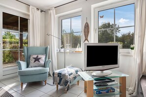 A living room with a blue armchair, a flat-screen TV on a glass table, and a view of greenery outside.