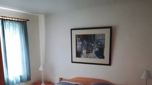Un dormitori amb un llit de fusta, una tauleta de nit amb una làmpada, un quadre abstracte emmarcat a la paret i una finestra amb cortines.