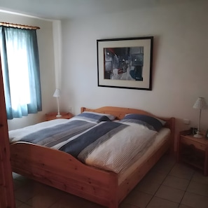 Ein Schlafzimmer mit einem hölzernen Bett, einem Nachttisch mit Lampe, einem gerahmten abstrakten Gemälde an der Wand und einem Fenster mit Vorhängen.