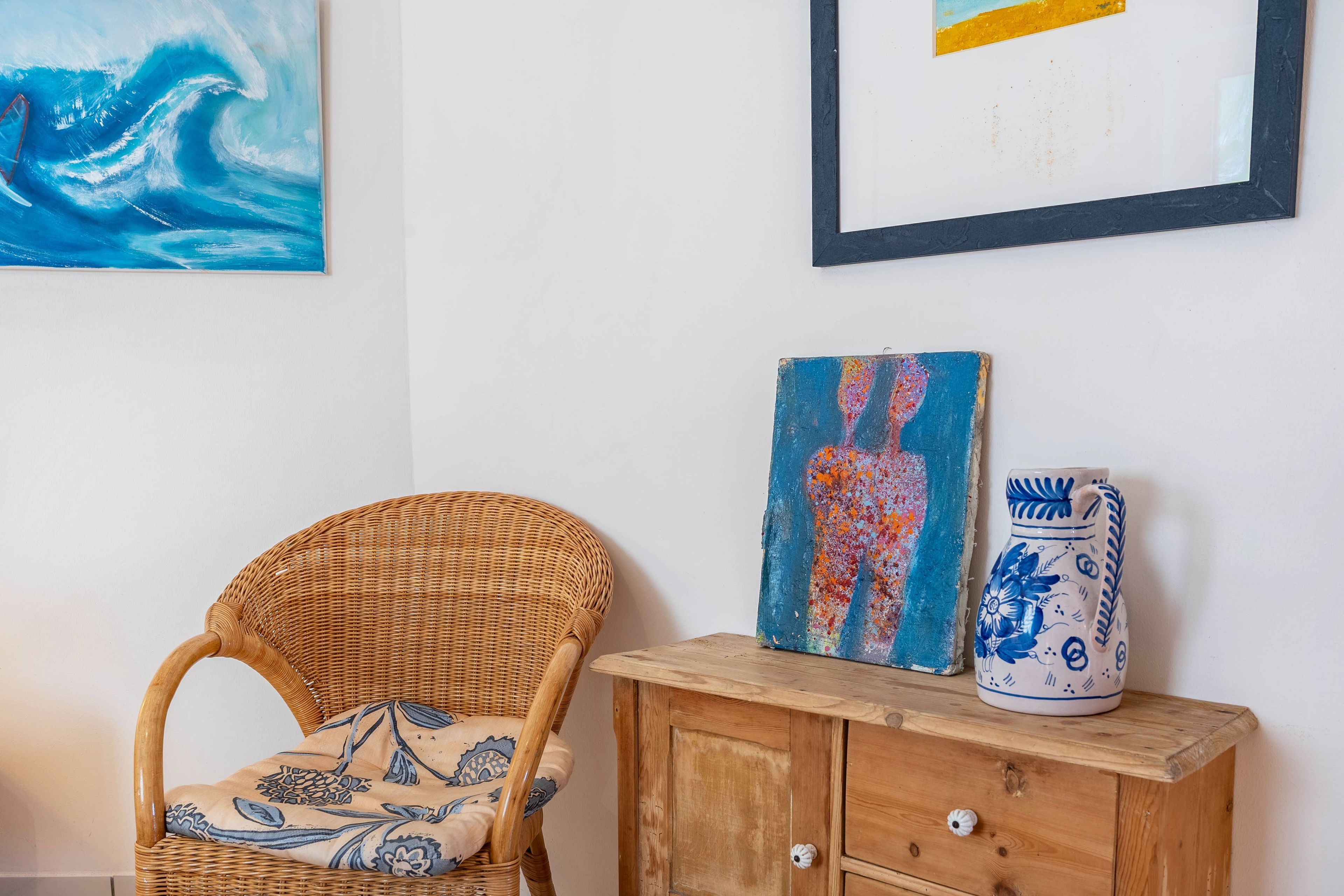 A wicker chair with a patterned cushion, a wooden cabinet with a vase, and two artworks on the wall.