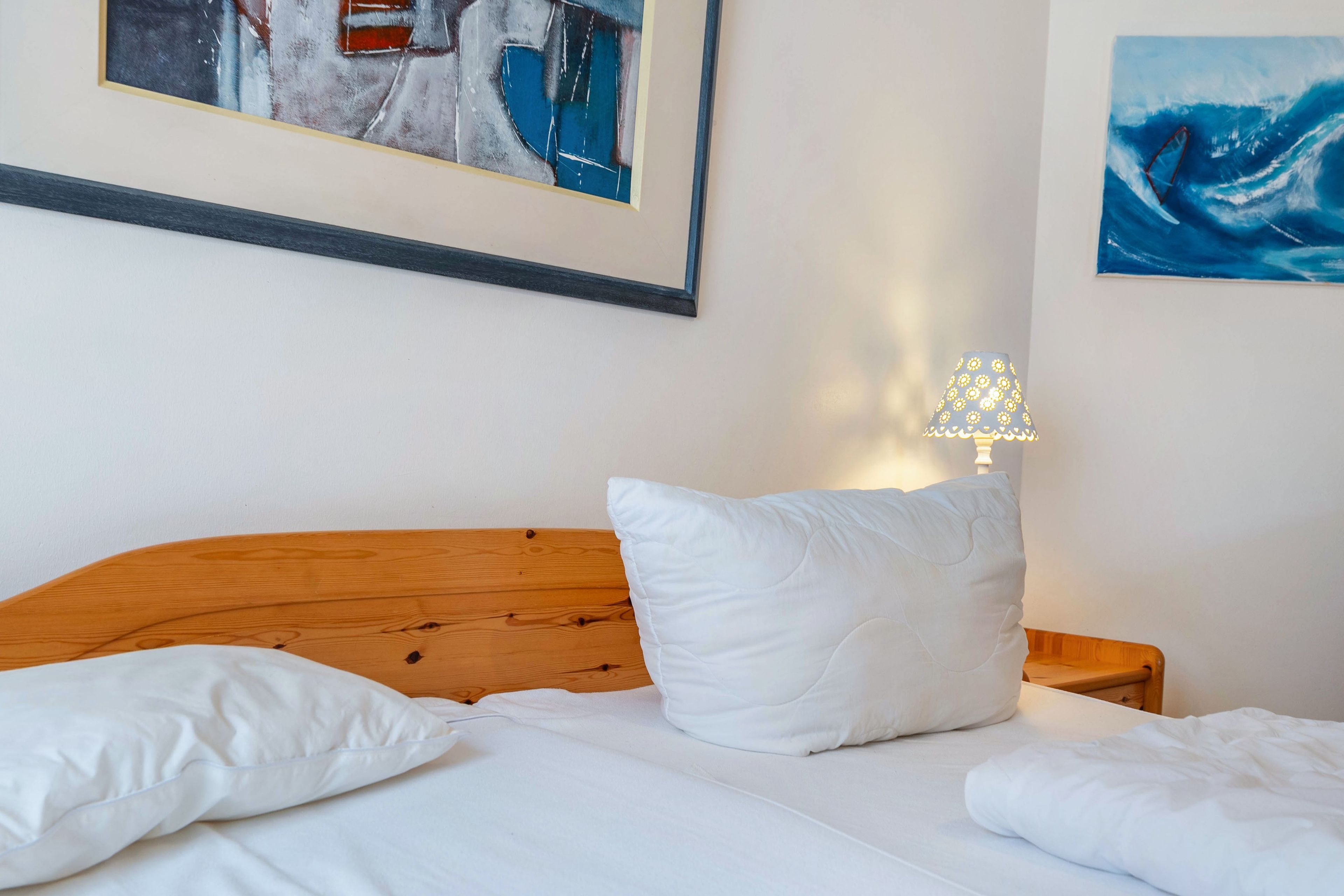 Une chambre avec une tête de lit en bois, une literie blanche et deux oreillers. Un tableau abstrait encadré est accroché au-dessus du lit, et une lampe se trouve sur la table de chevet.