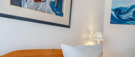 Un dormitorio con cabecera de madera, ropa de cama blanca y dos almohadas. Hay un cuadro abstracto enmarcado sobre la cama y una lámpara en la mesita de noche.