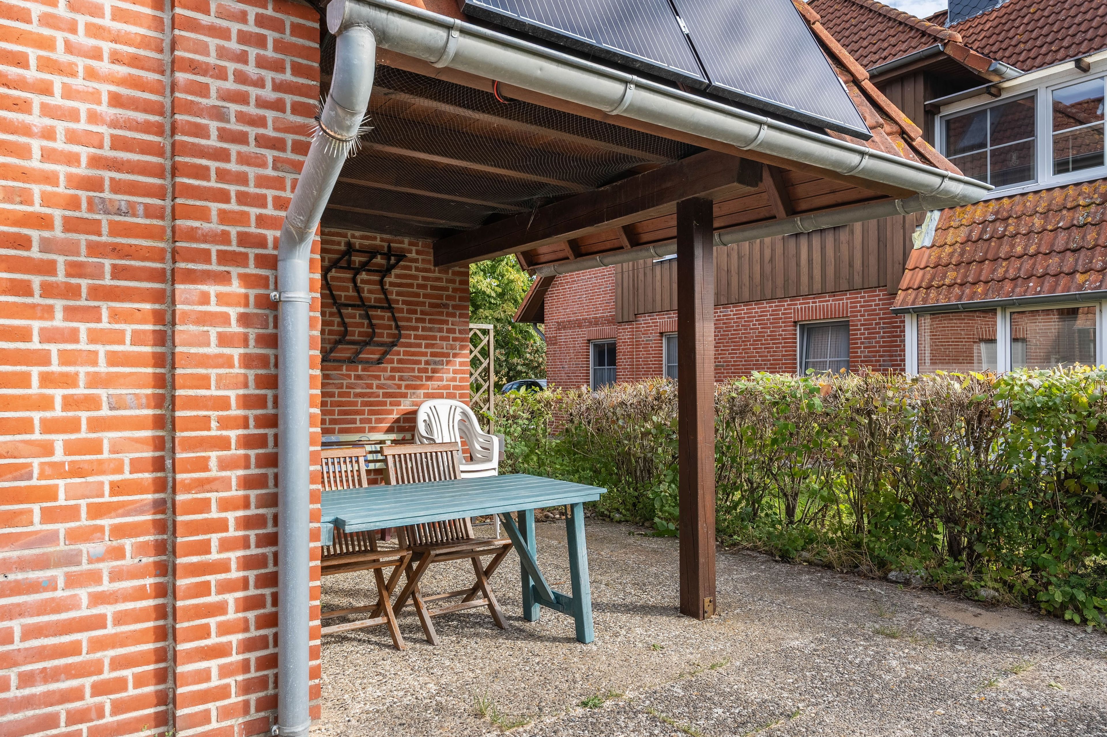 Een overdekt terras met een tafel en stoelen, een bakstenen muur en een haag.